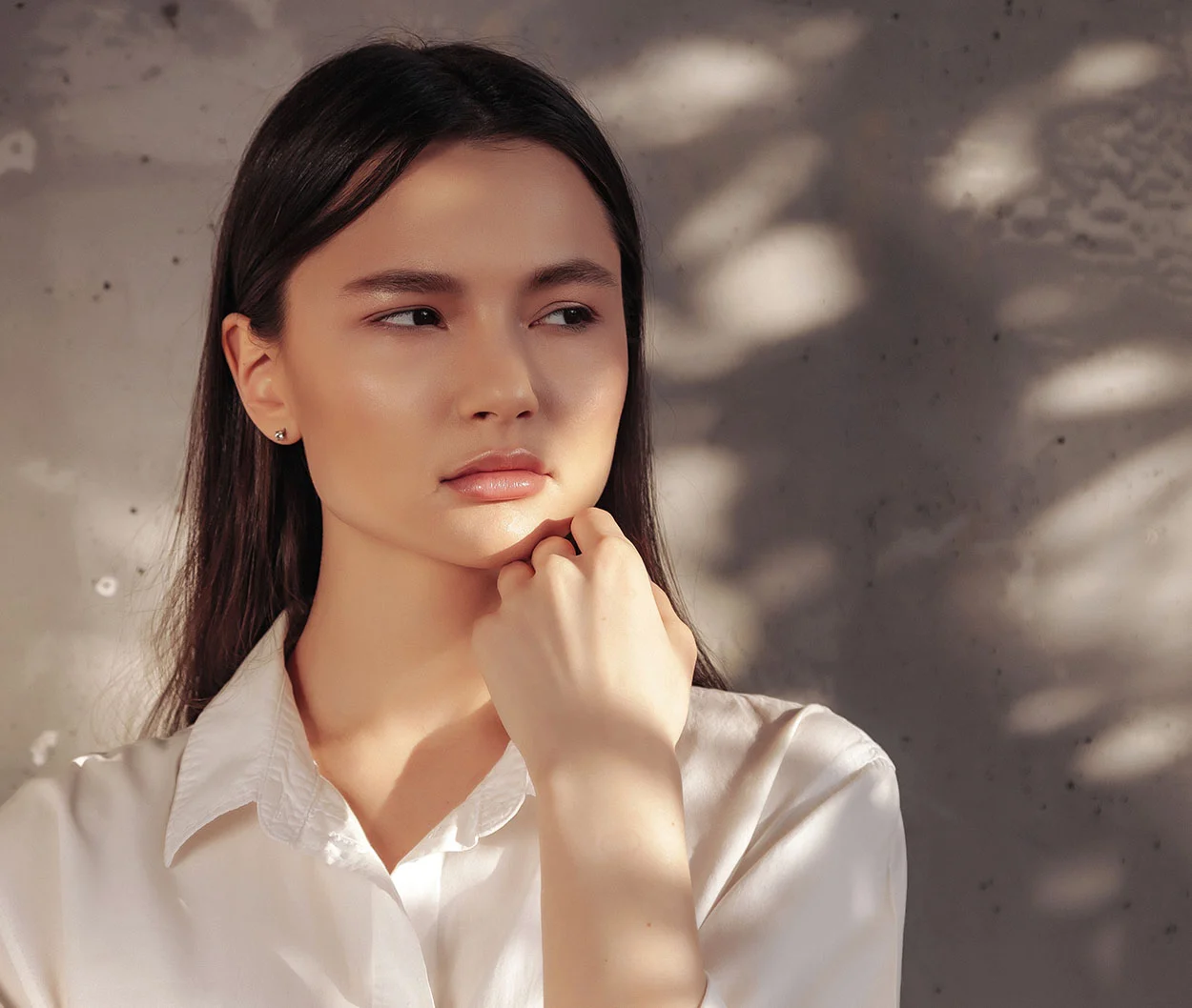 A woman standing against a textured wall, wearing a white shirt, her dark hair falling over her shoulders, with a thoughtful and pensive expression - Chemical Peels in Marietta, GA