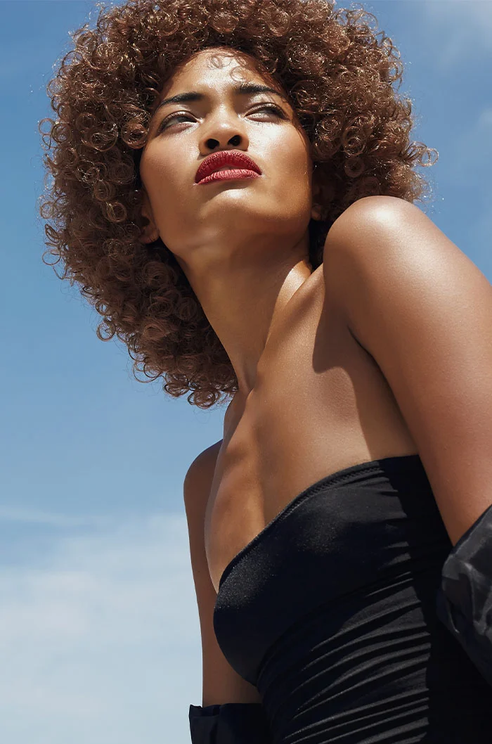 A woman with voluminous curly hair, wearing a strapless black top, posing under the bright sunlight with a confident and fierce expression - Dermal Fillers in Marietta, GA